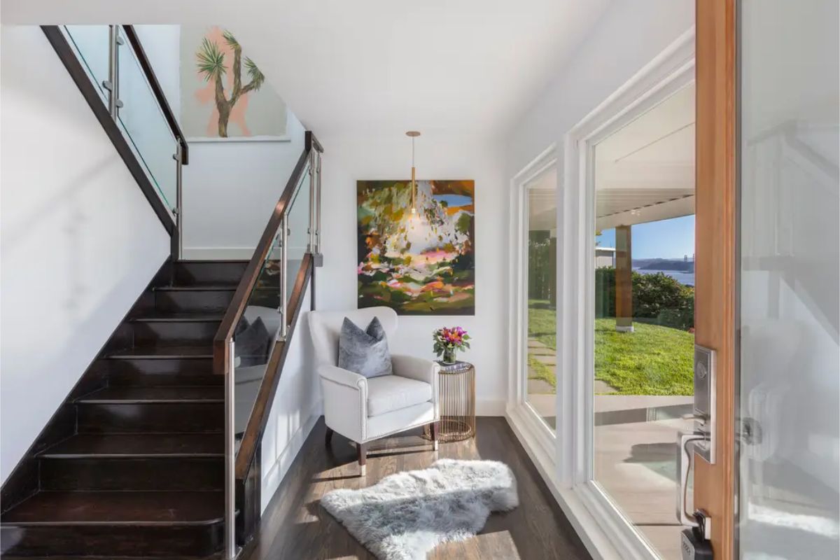 Bright entryway with a staircase, glass railing, and a cozy seating nook with natural light pouring in. 
