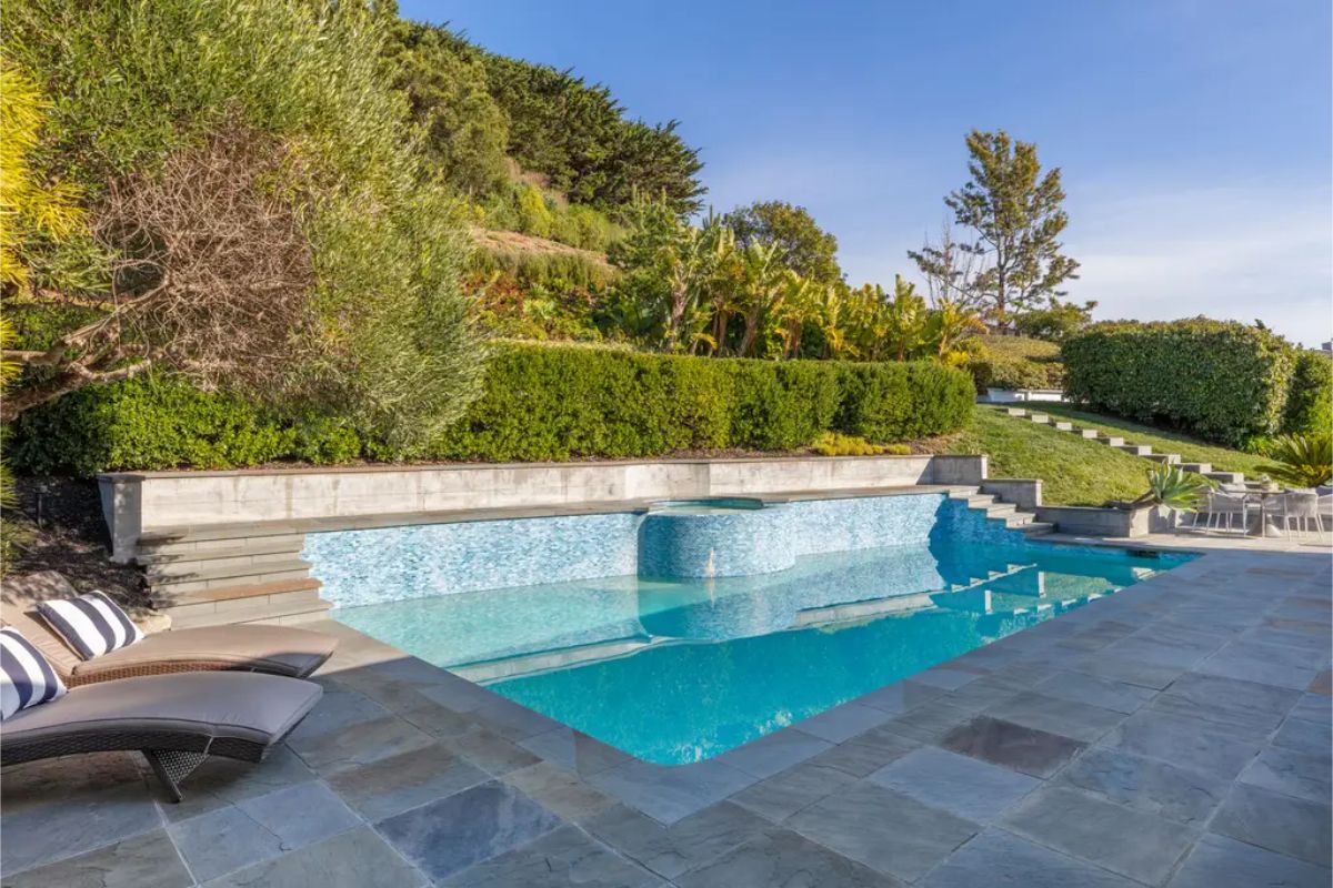 Serene outdoor pool with a built-in spa, surrounded by lush landscaping and stone-paved lounging areas. 