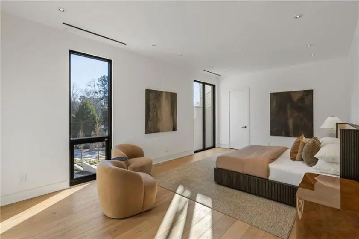 Minimalist bedroom features earthy tones, tall windows, and cozy lounge chairs in a bright, serene setting. 
