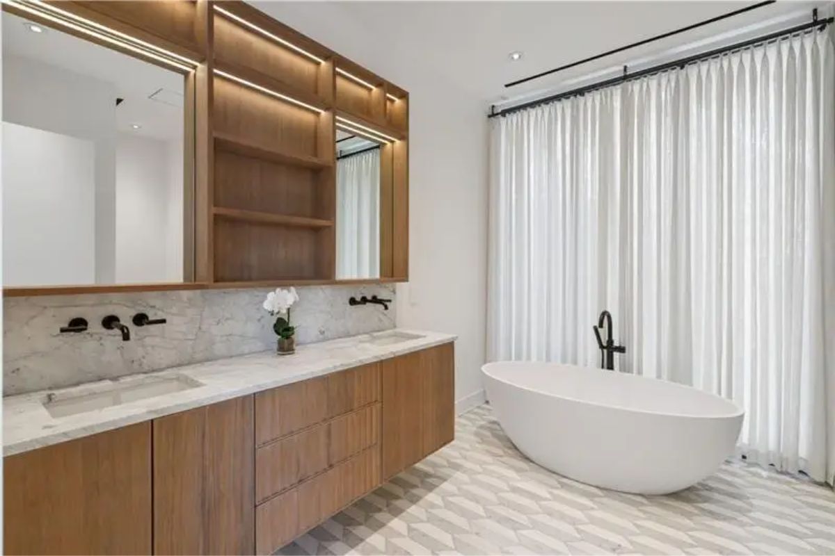 Light-filled bathroom boasts a freestanding tub, dual sinks with wood cabinetry, and geometric patterned flooring. 