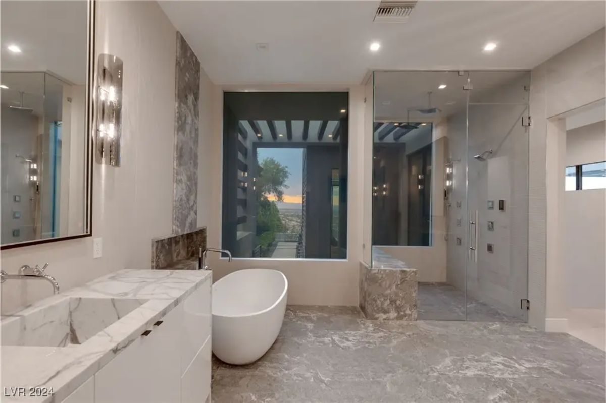 A luxurious bathroom features a freestanding white bathtub, dual vanities, and a large glass-enclosed shower with a scenic outdoor view. 