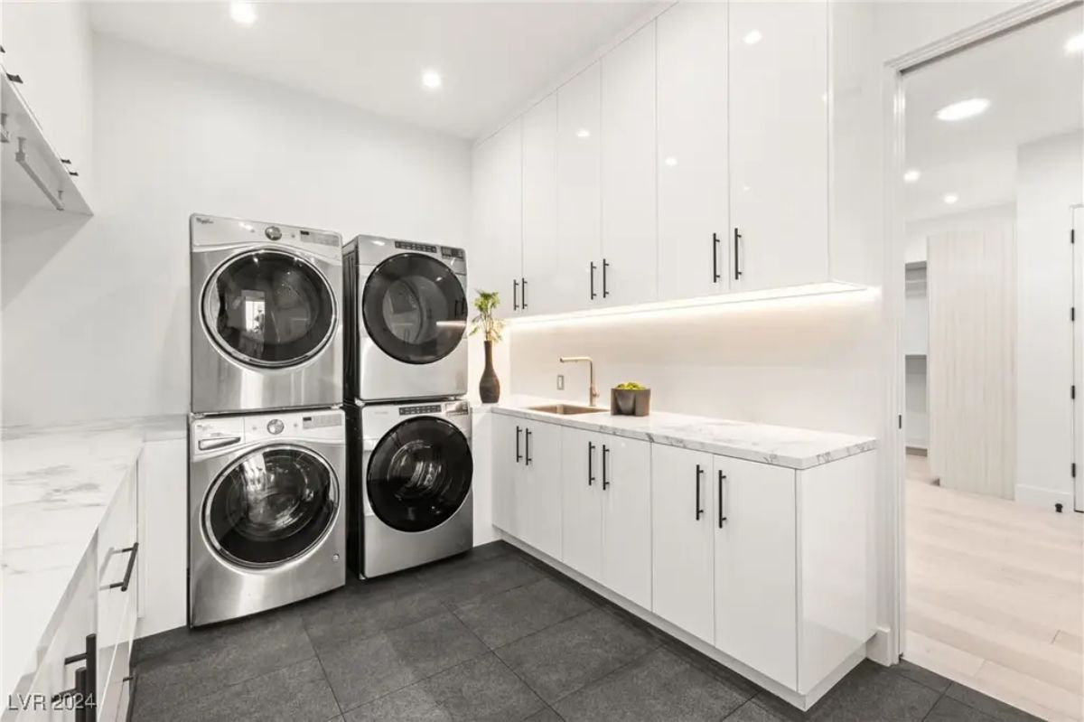 A modern laundry room is equipped with two stacked washers and dryers, sleek white cabinetry, and a spacious countertop. 