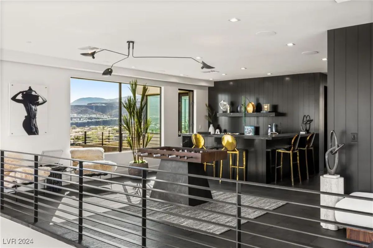 A stylish game room includes a foosball table, a bar area with high stools, and modern décor featuring large windows. 