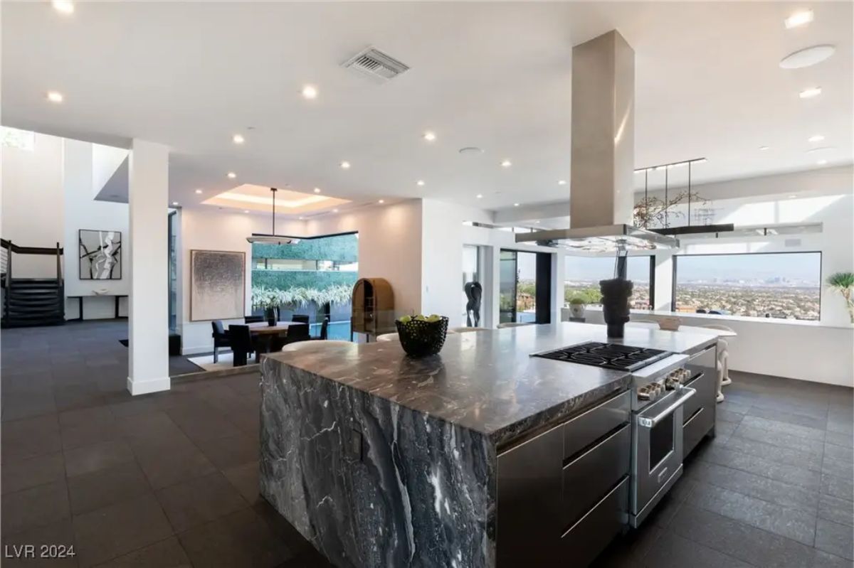 A spacious kitchen with a massive island featuring dark marble, stainless steel appliances, and an open-concept layout connecting to the dining and living areas. 
