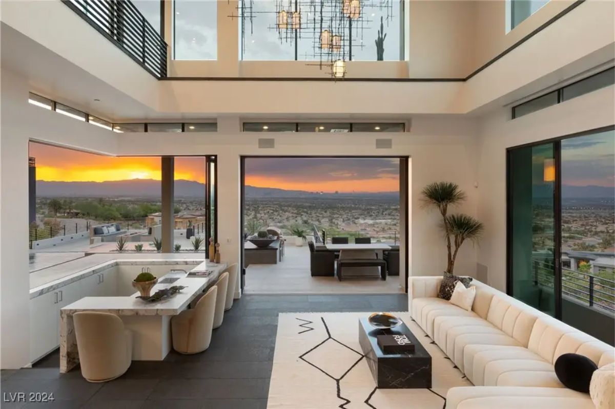 A luxurious living room with a white sectional sofa, high ceilings, and floor-to-ceiling windows that frame a stunning sunset view. 