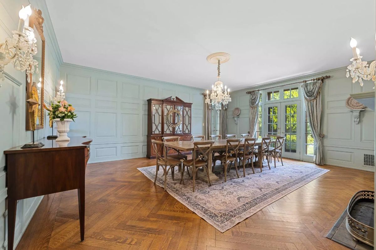 Long wooden table is surrounded by ten chairs on a large patterned rug.