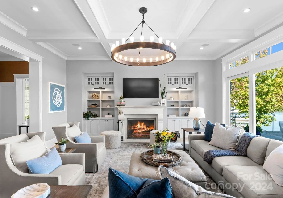 Cozy living room centers around a fireplace and coffered ceiling, with views of the water beyond. 