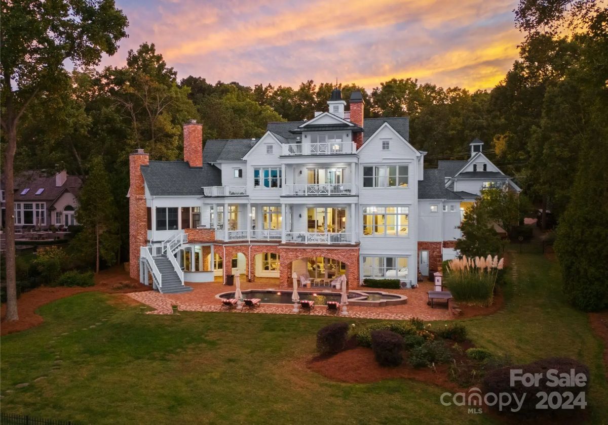 Three-story lakeside estate with brick and white siding details, multiple balconies, and a pool patio set against a vibrant sunset backdrop.
