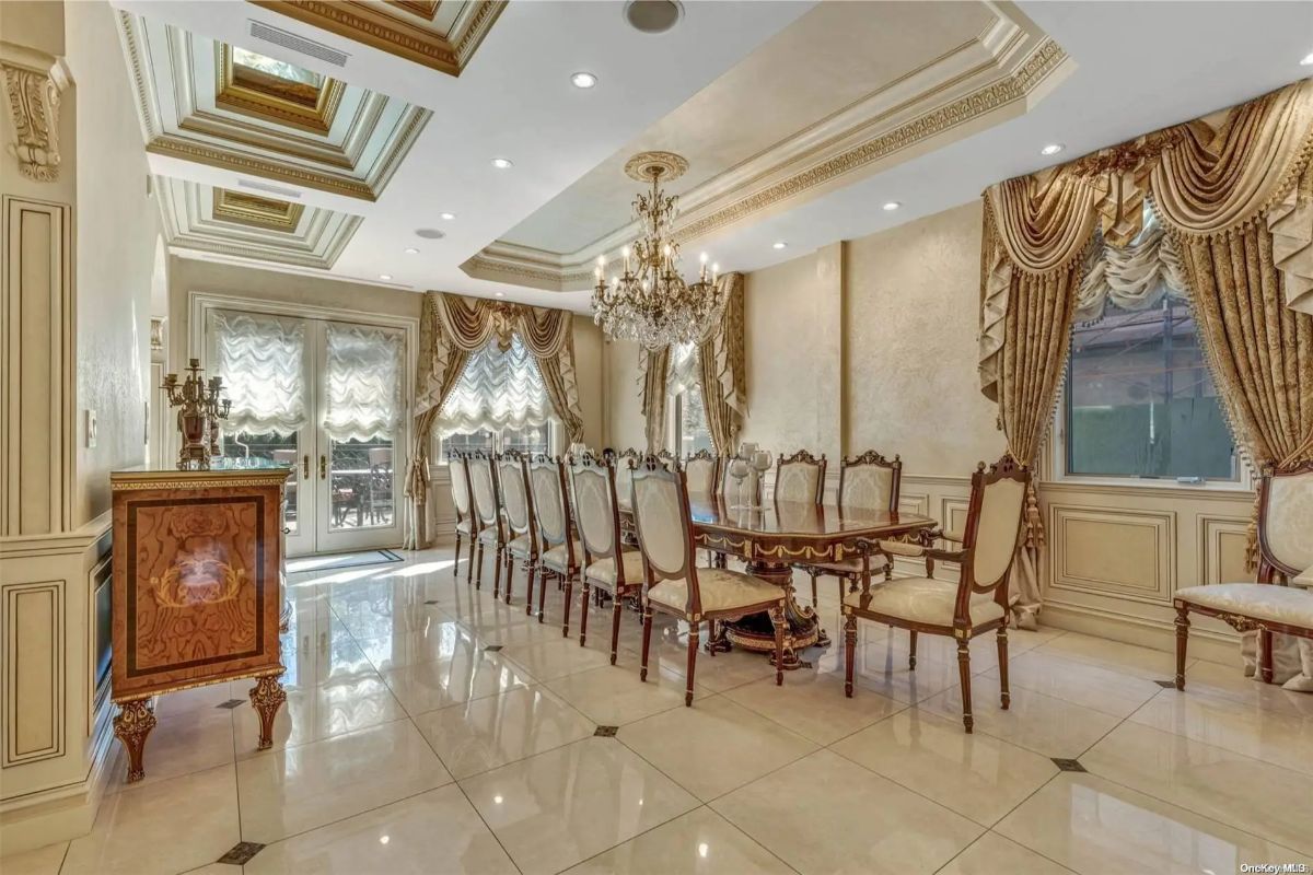 Long dining table is surrounded by high-back carved chairs and positioned beneath a crystal chandelier.