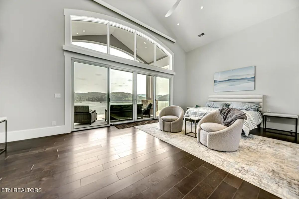 Luxurious bedroom with a vaulted ceiling, large arched windows, and sliding glass doors leading to a private balcony with lake views. 