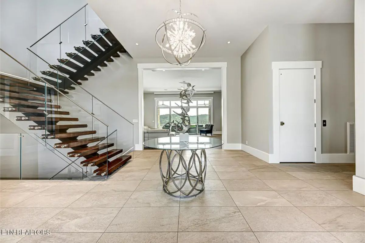 Sleek entryway featuring a floating staircase with glass railings, a contemporary chandelier, and an elegant round table centerpiece. 