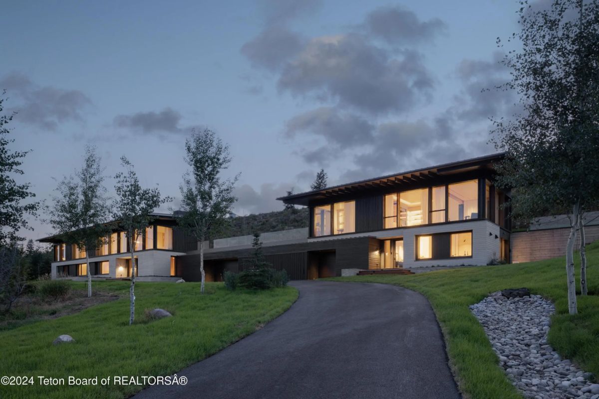 Evening view of a sleek contemporary residence with glowing windows and a private drive surrounded by trees. 