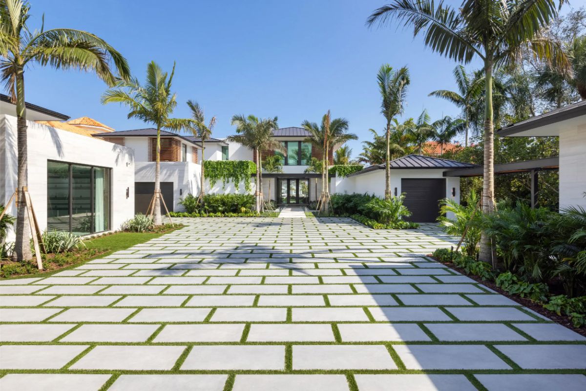 Wide paver driveway leads to a contemporary mansion framed by tall palm trees. 