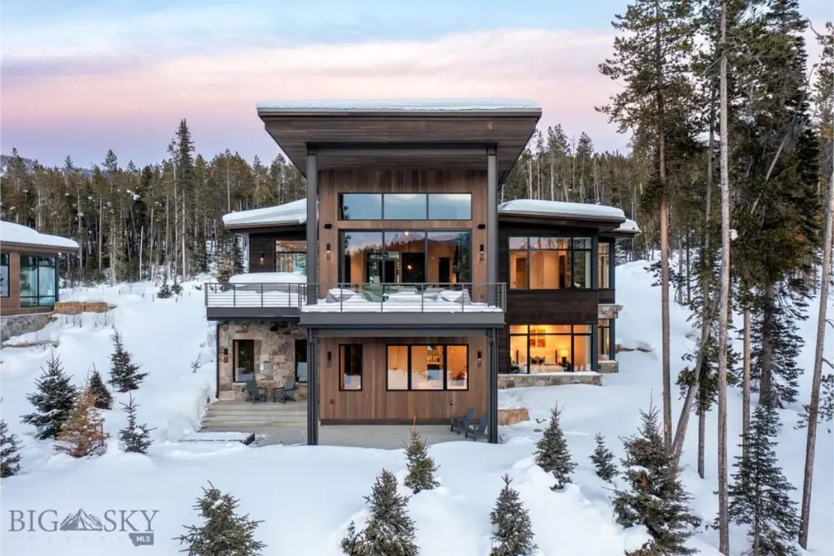Modern mountain home with floor-to-ceiling windows is nestled in a snowy forest, showcasing a sleek architectural design.