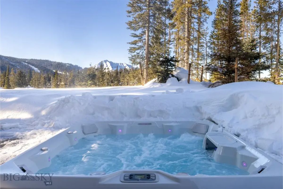 Outdoor hot tub surrounded by deep snow overlooks a serene forested landscape with mountains in the background.