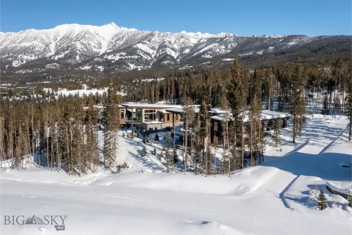 Expansive aerial view of a luxurious snow-covered estate is set against a backdrop of towering mountains and dense evergreen trees.