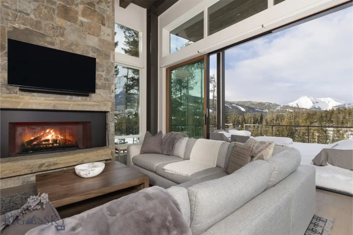 Expansive glass doors open to a snowy mountain view, complementing the contemporary fireplace and neutral-toned furniture.