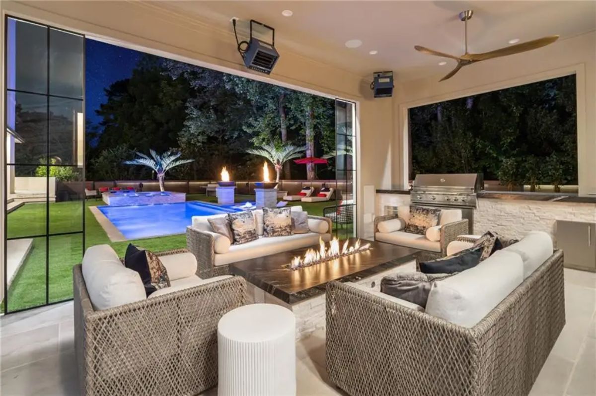 Covered outdoor lounge with a sleek fire pit, cozy wicker seating, and full view of the illuminated pool and landscaped yard at night. 