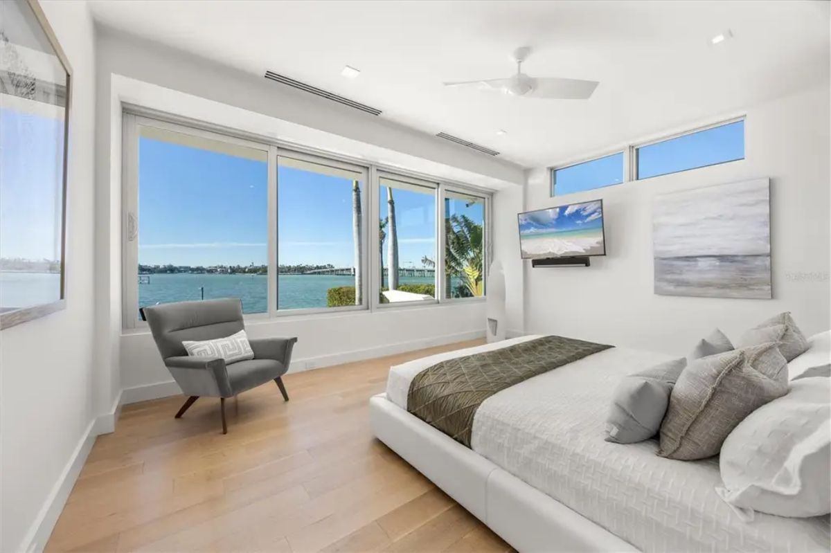 Serene bedroom with large windows offering a stunning waterfront view, minimalist decor, and a cozy reading chair. 