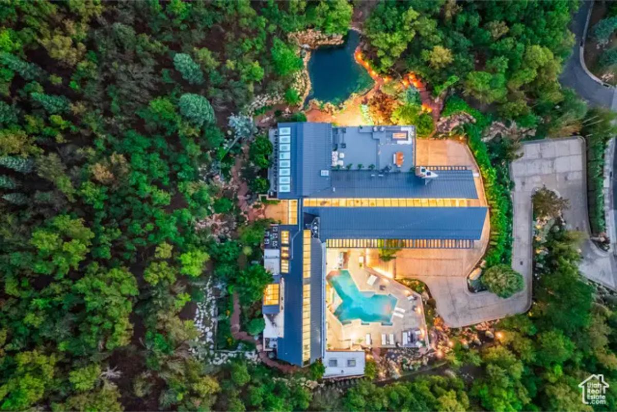 An aerial shot of a modern mansion nestled in a lush green forest, showcasing an expansive property with a pool and landscaped grounds. 