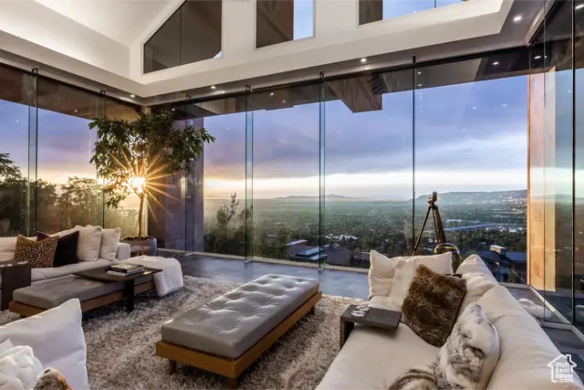 Living room with a floor-to-ceiling glass wall, offering an unobstructed panoramic view of the city and sunset. 