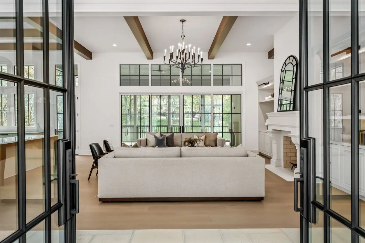 View into the airy living room with exposed beams, chandelier, and large paned windows letting in natural light. 