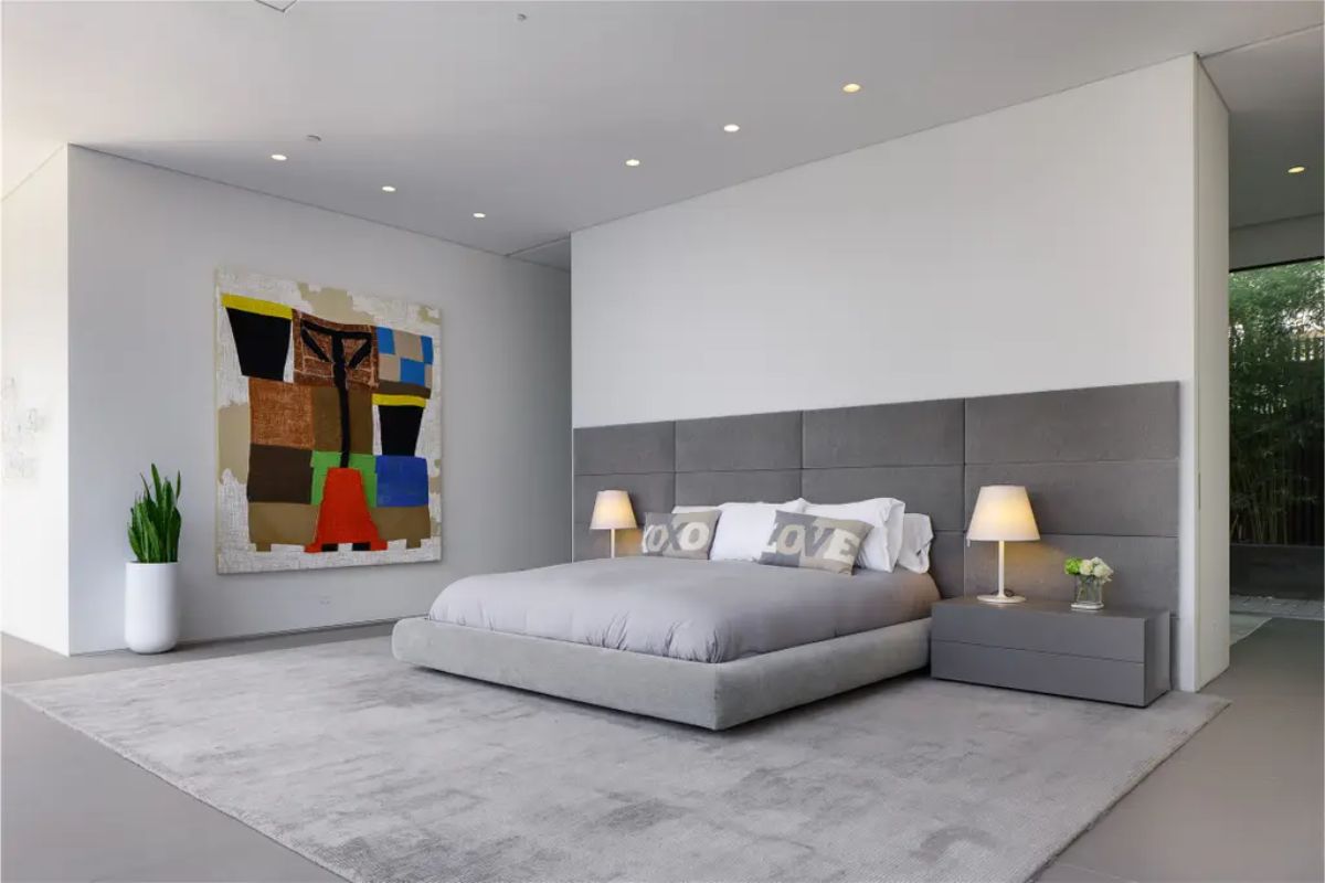 A spacious bedroom with a gray upholstered headboard, ambient lighting, and a bold abstract painting. 