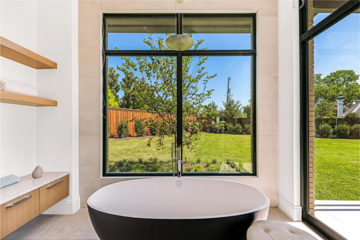 Freestanding black and white bathtub positioned in front of floor-to-ceiling windows overlooking a lush backyard. 