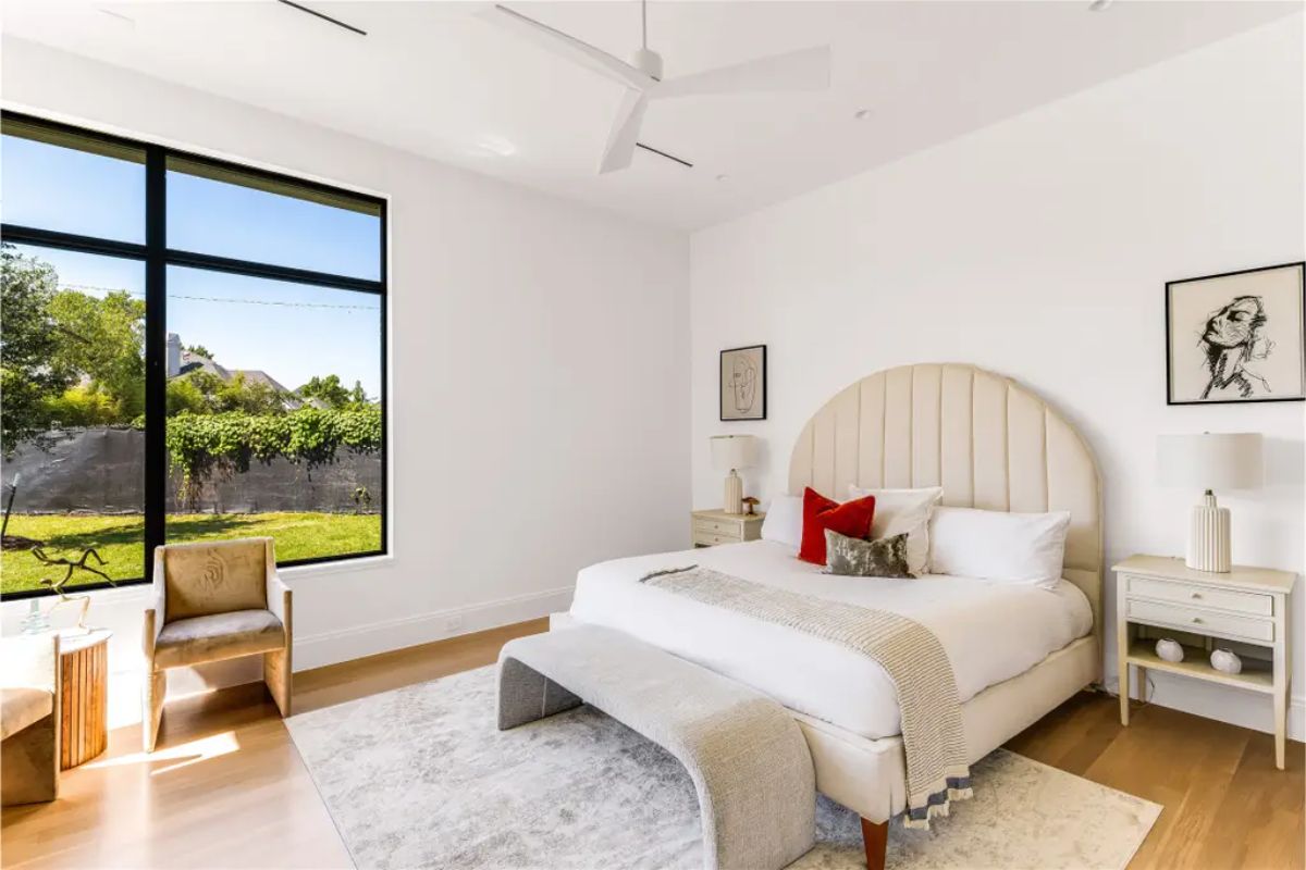 Cozy guest bedroom featuring a curved upholstered headboard, neutral tones, and large windows with a backyard view. 