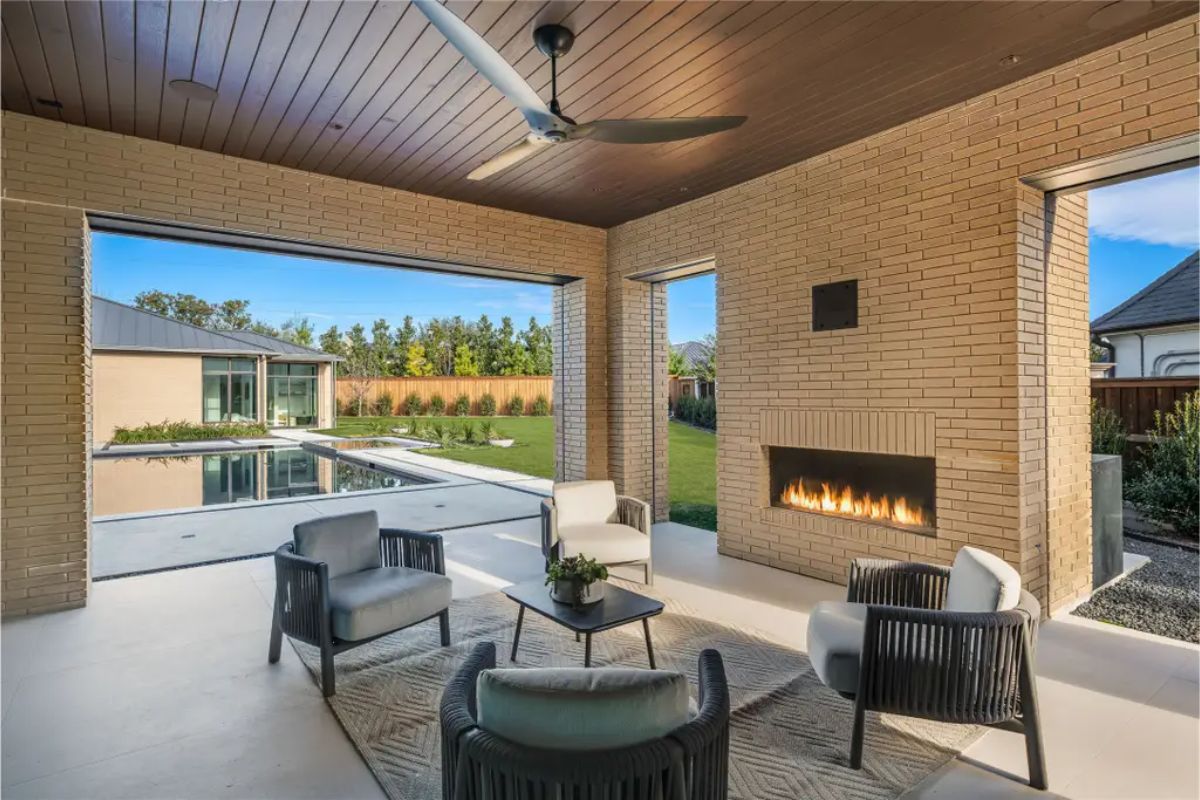 Covered outdoor lounge area with a fireplace, seating arrangement, and a wooden ceiling with a ceiling fan. 