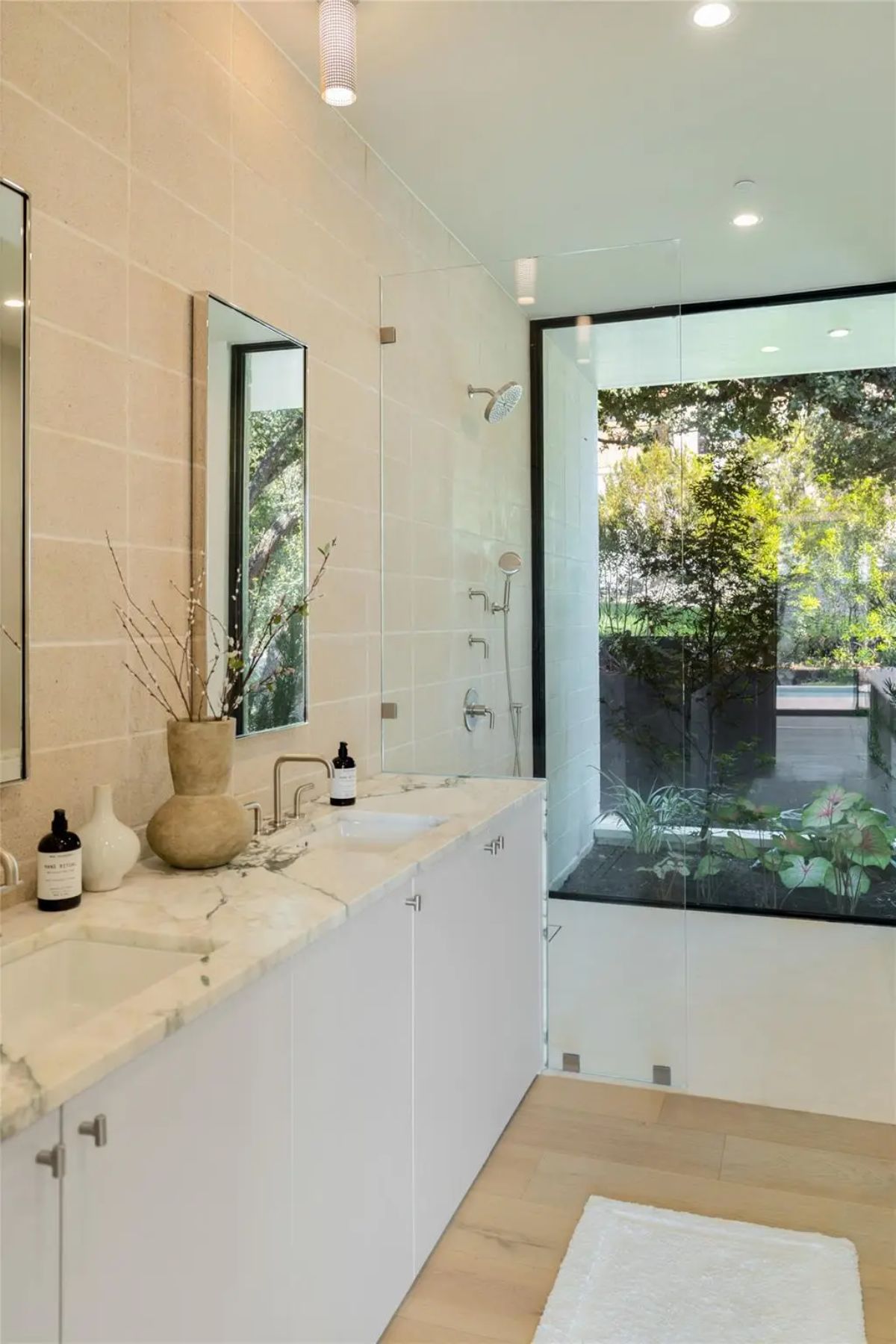 Luxurious bathroom with a dual vanity, marble countertops, and a glass-enclosed shower overlooking lush greenery. 