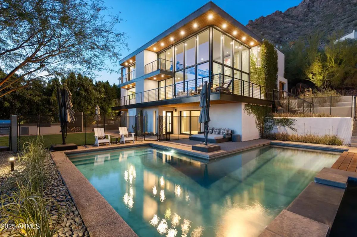 Modern three-story home with extensive glass walls, warm exterior lighting, and a luxurious pool reflecting the house at dusk.