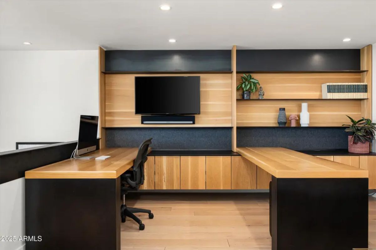 Sleek home office with built-in wooden and black shelving, dual workstations, and a mounted TV for a modern workspace. 