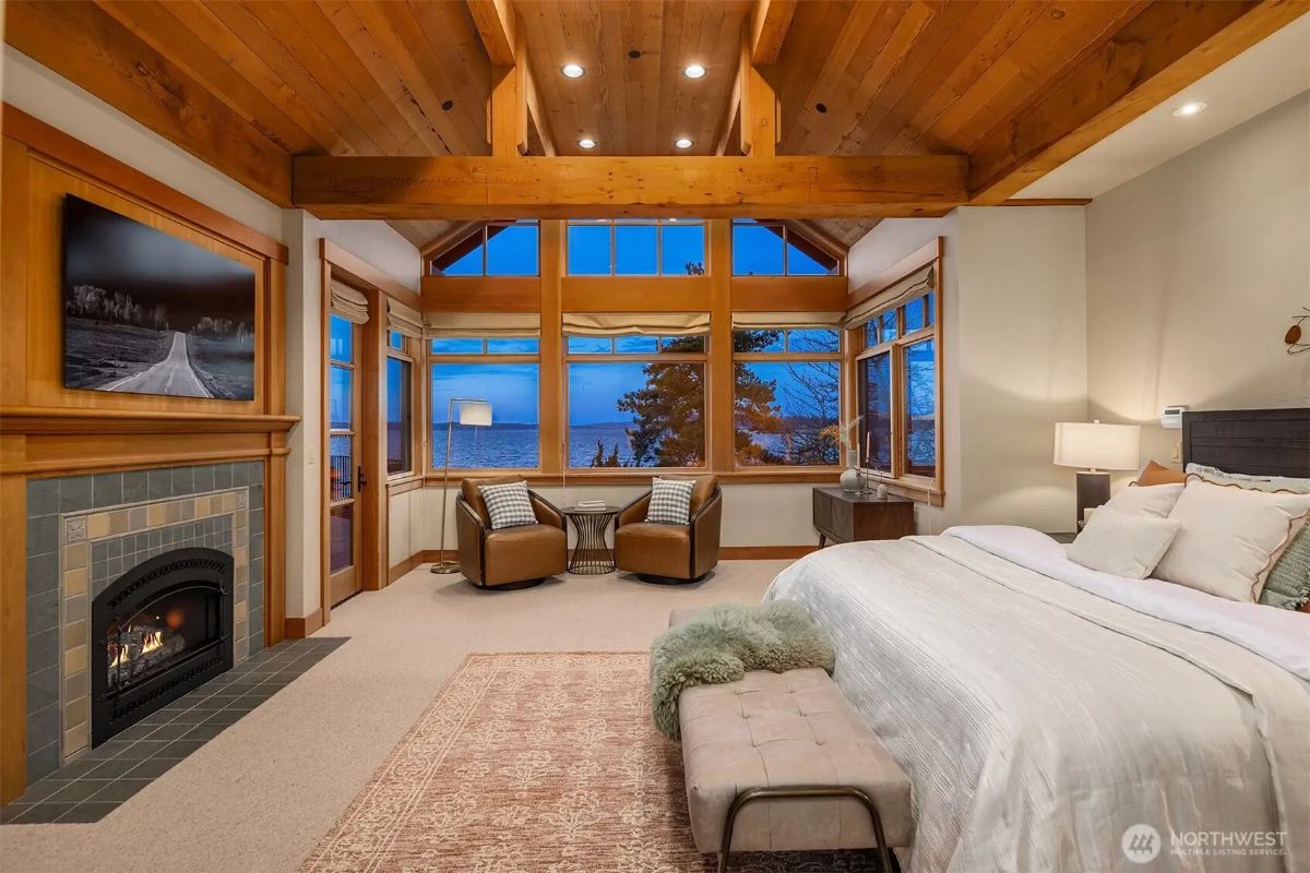 Luxurious primary bedroom with vaulted wood ceilings, a fireplace, and panoramic lake views through large windows. 