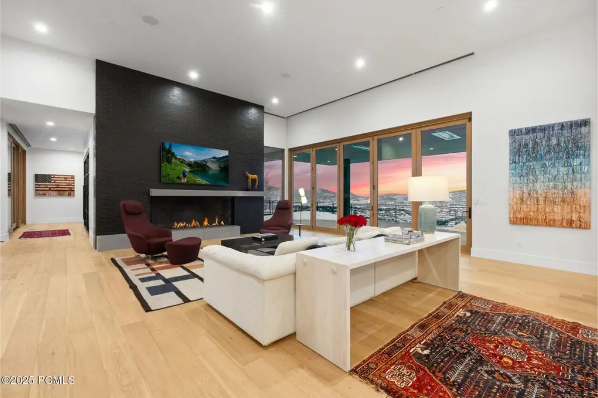 Cozy living room boasts a fireplace, dark accent walls, and modern seating, with a breathtaking sunset view through floor-to-ceiling windows. 