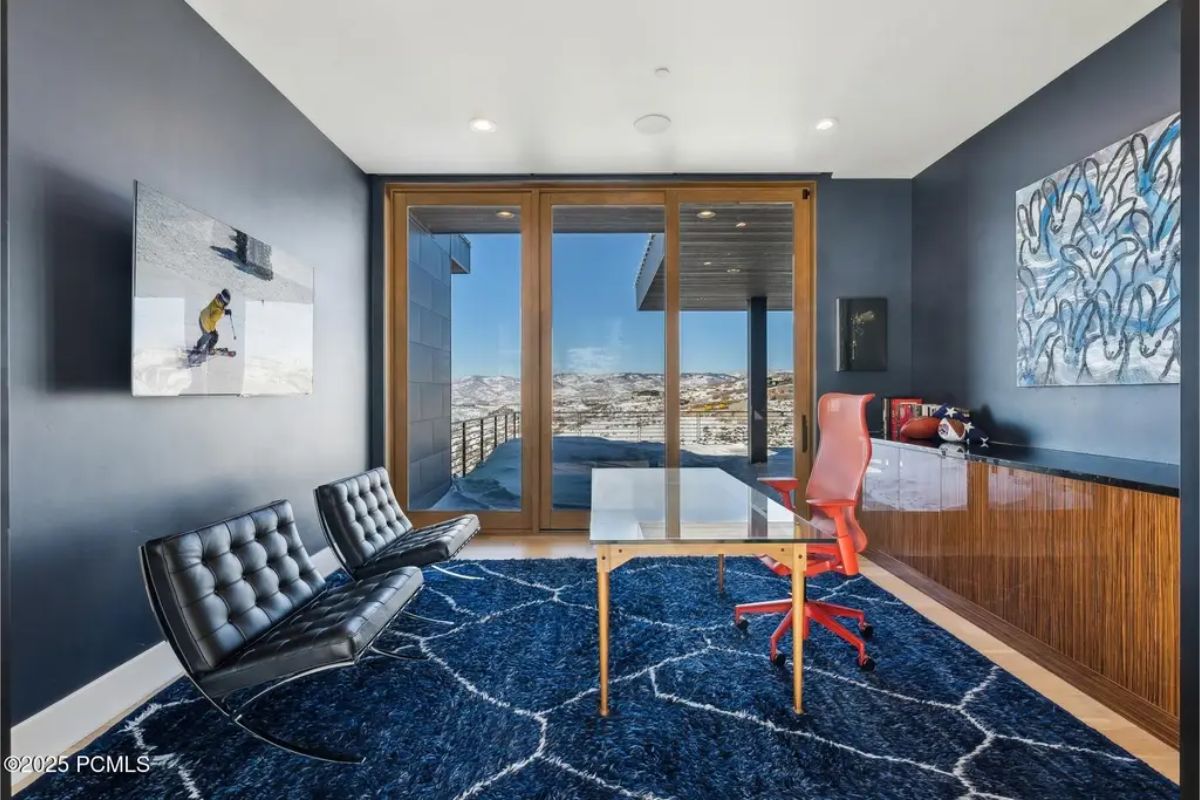 Home office with a contemporary blue theme, a glass desk, and large windows offering panoramic mountain views. 