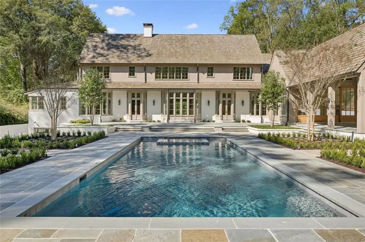 Rectangular pool and spa with a stone patio centered in front of a grand modern farmhouse. 