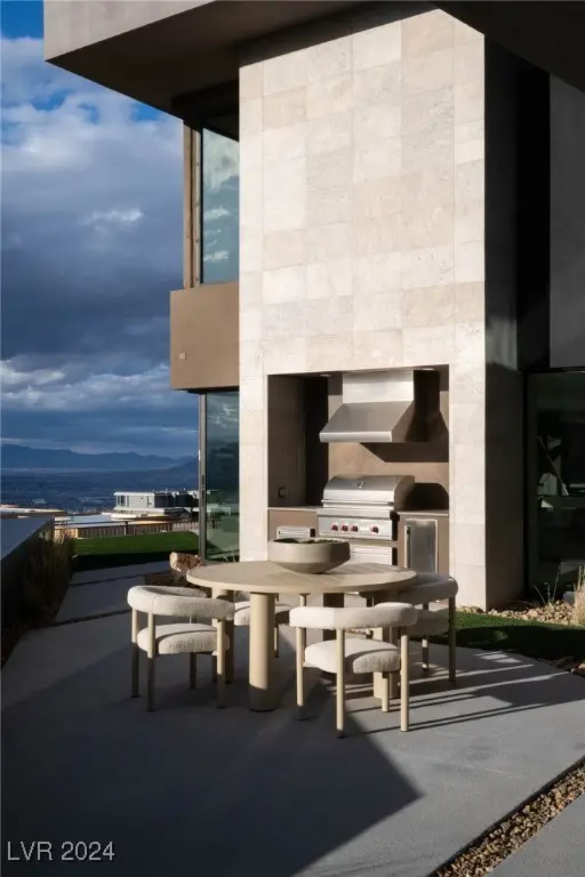 Outdoor dining area with built-in grill and round table beside a stone exterior wall.