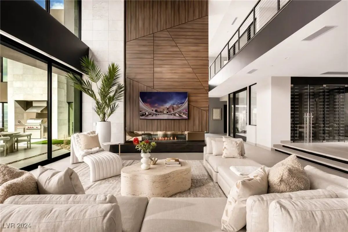 Living room with double-height ceilings, wood-paneled fireplace wall, and access to patio and pool.
