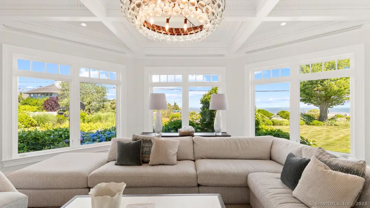 Window-wrapped sitting area with large sectional, coffered ceiling, and lush garden and water views. 