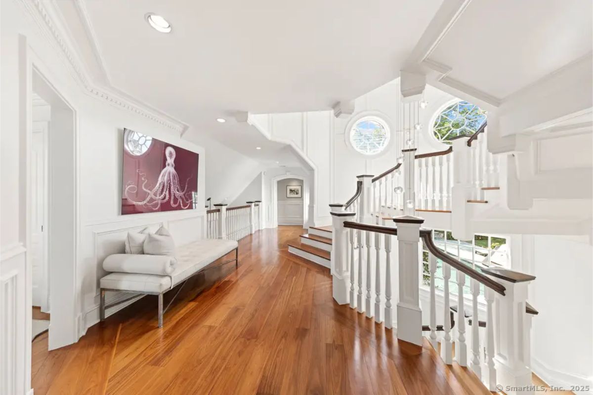 Grand hallway with curved staircase, circular windows, hardwood floors, and contemporary wall art. 