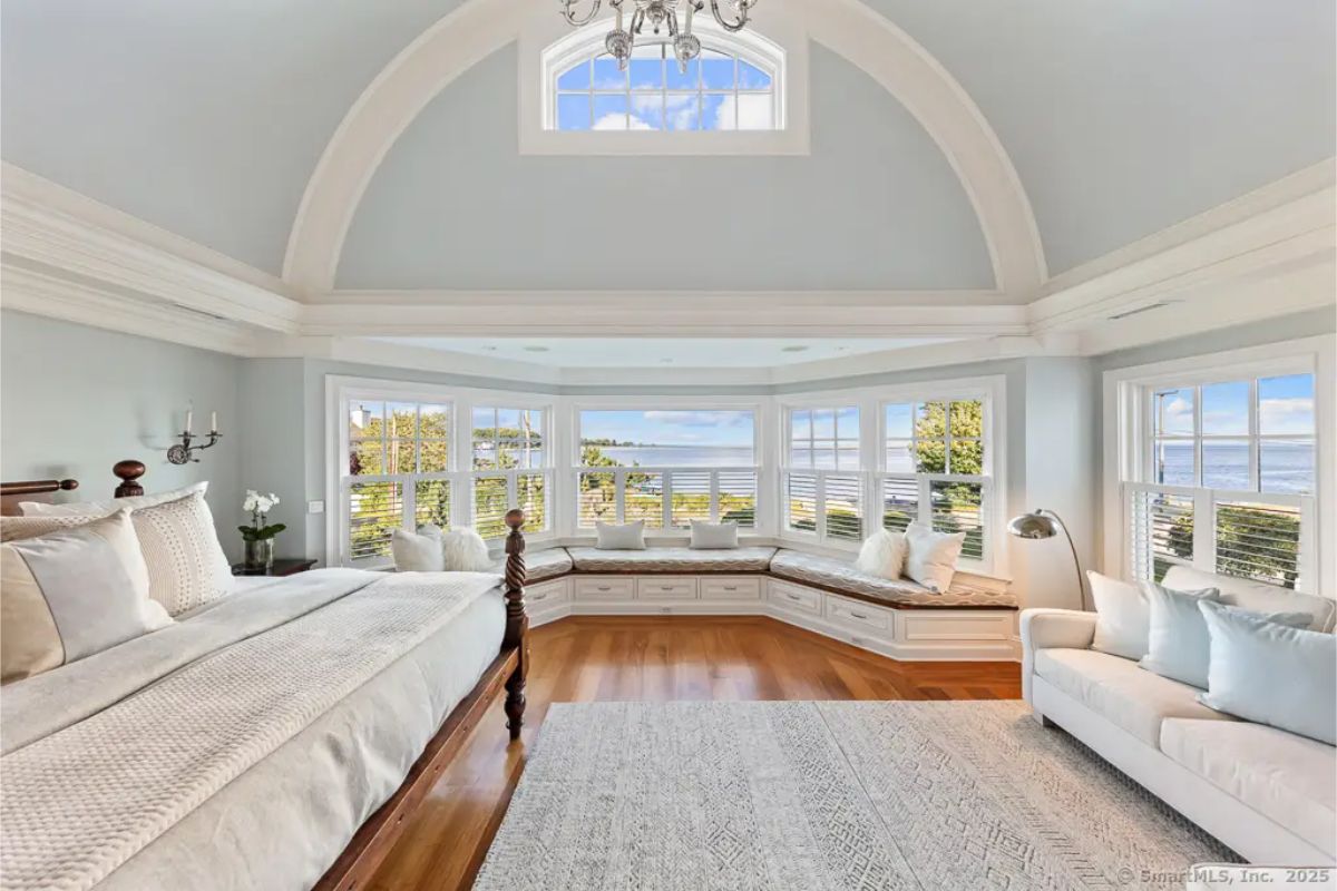 Vaulted primary bedroom with arched ceiling, panoramic water views, built-in window seating, and traditional furnishings. 