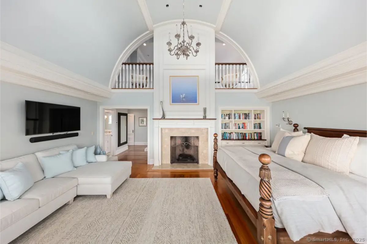Vaulted bedroom with fireplace, built-in bookshelves, double-height ceiling, and upper interior balcony. 