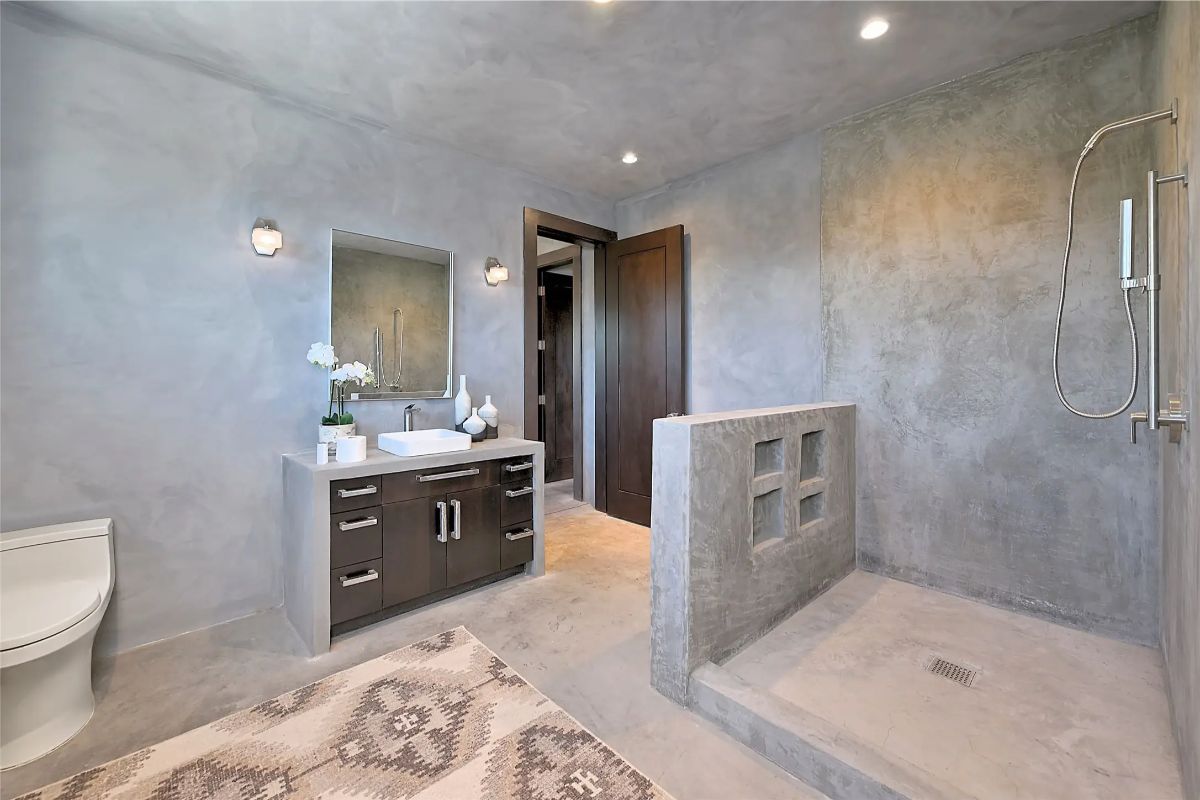 Minimalist concrete bathroom with open shower, wall sconces, and modern vanity. 
