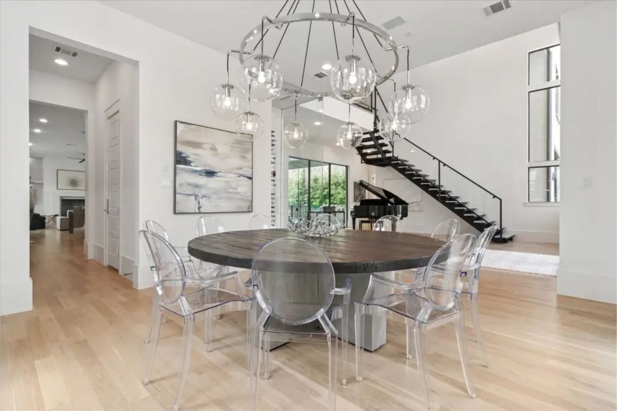 Dining area with round table, clear acrylic chairs, and modern chandelier beneath floating staircase. 