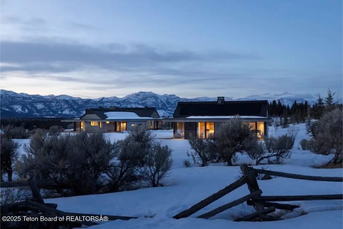 Evening view of snow-covered home with soft interior lighting and distant mountain backdrop. 