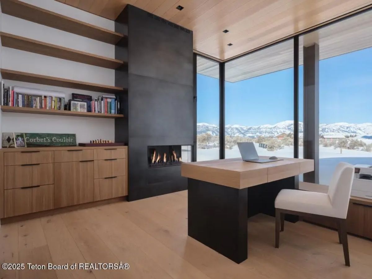 Office with built-in shelves, modern fireplace, and desk facing floor-to-ceiling windows with snow-covered scenery. 