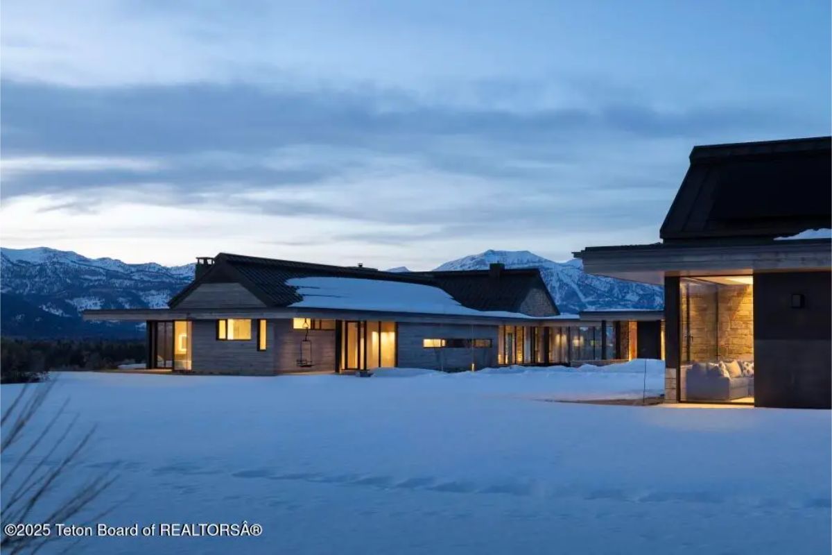 Twilight exterior of modern mountain residence with glowing windows and expansive snowy terrain. 