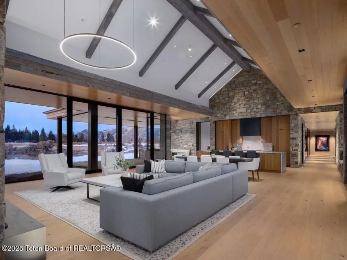 Open-concept living space with vaulted ceiling, exposed beams, stone accents, and floor-to-ceiling windows. 