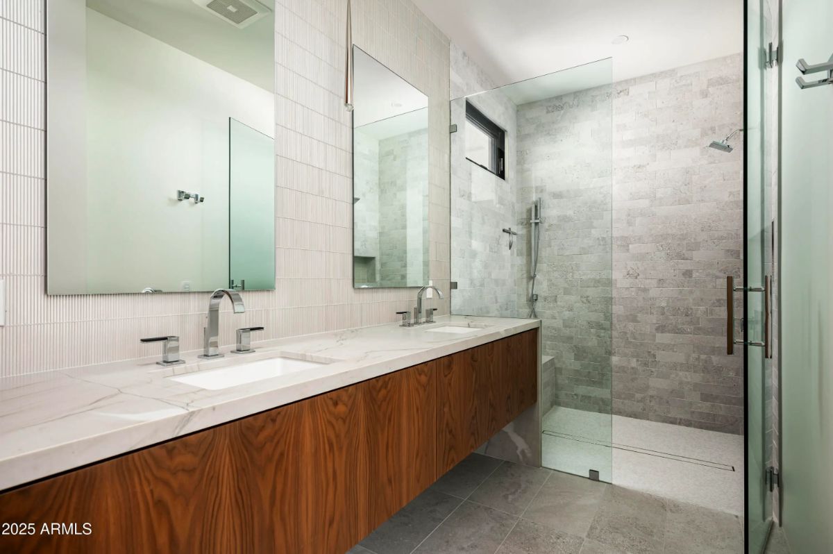 Dual-sink bathroom with long wood vanity and glass-enclosed stone-tile shower. 